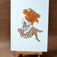 Une femme lit un journal sous une immense feuille orange d’automne portant le titre « ALL WRONG? », œuvre originale d’Ana Jarén.