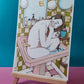 Une femme se recroqueville dans un lavabo rose entouré de miroirs, de flacons et d’objets de salle de bains, œuvre originale d’Ana Jarén.